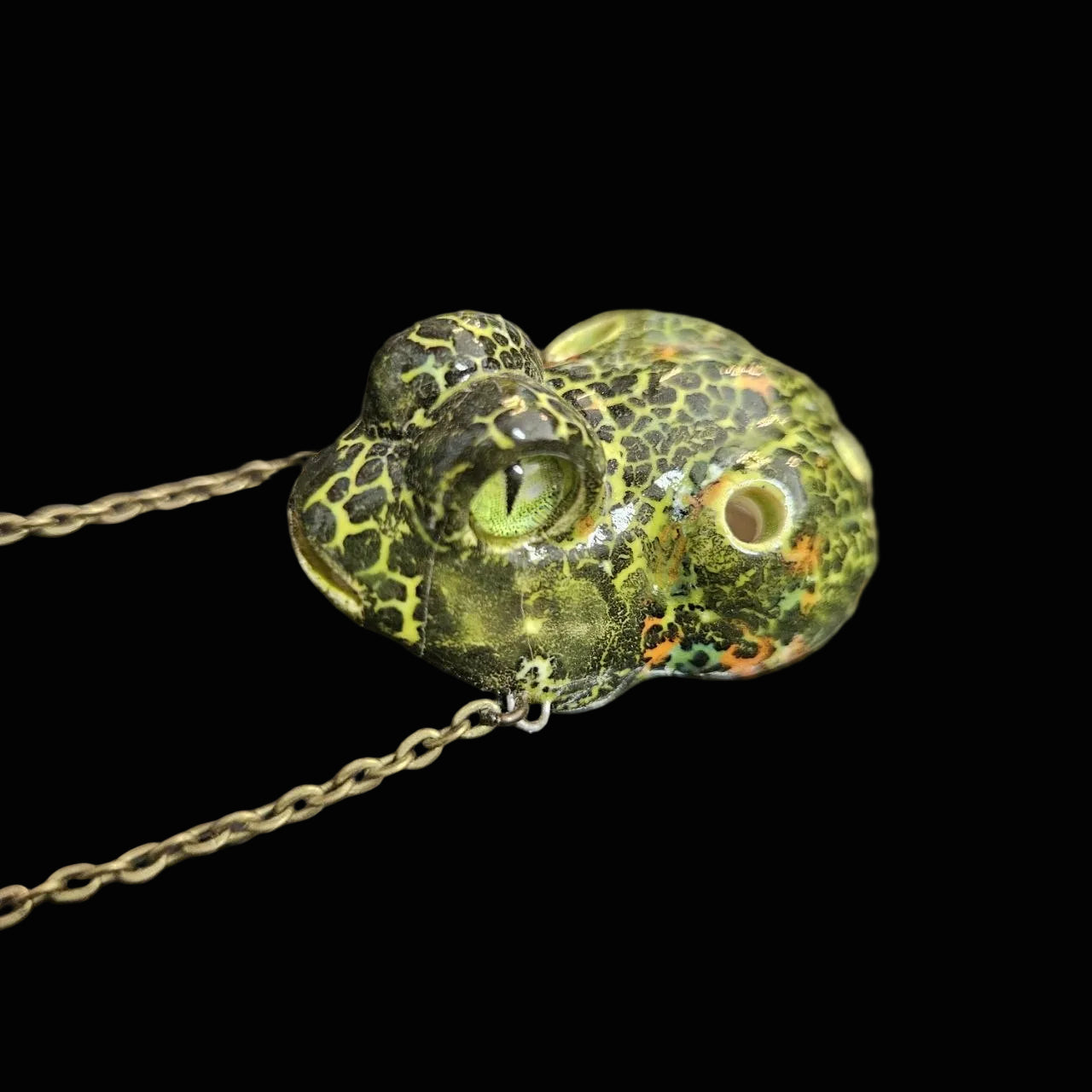 A close-up of a frog-shaped pendant necklace with a textured green and yellow mottled pattern, resting on a dark textured surface.