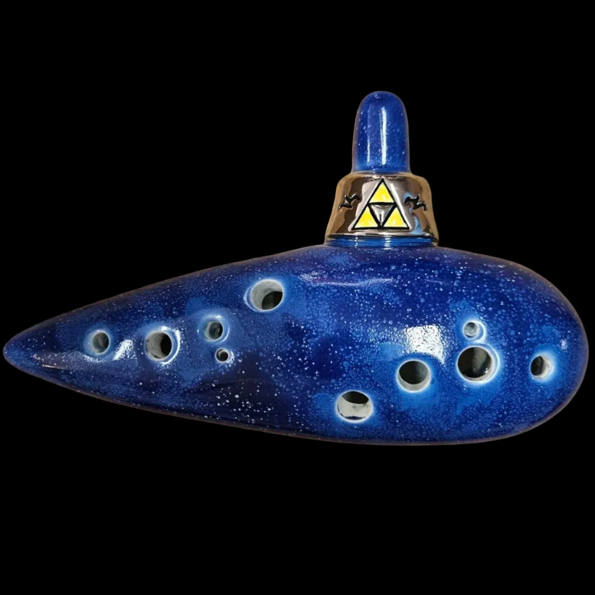 A handmade ceramic ocarina in deep blue with white speckles, featuring a silver metal neck with a yellow Triforce emblem, resting on a scratched wooden surface.