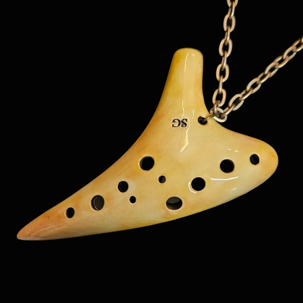 An ocarina-shaped pendant necklace in a glossy pale yellow ceramic, featuring multiple circular port holes and a gold-tone chain, displayed on a dark textured background.