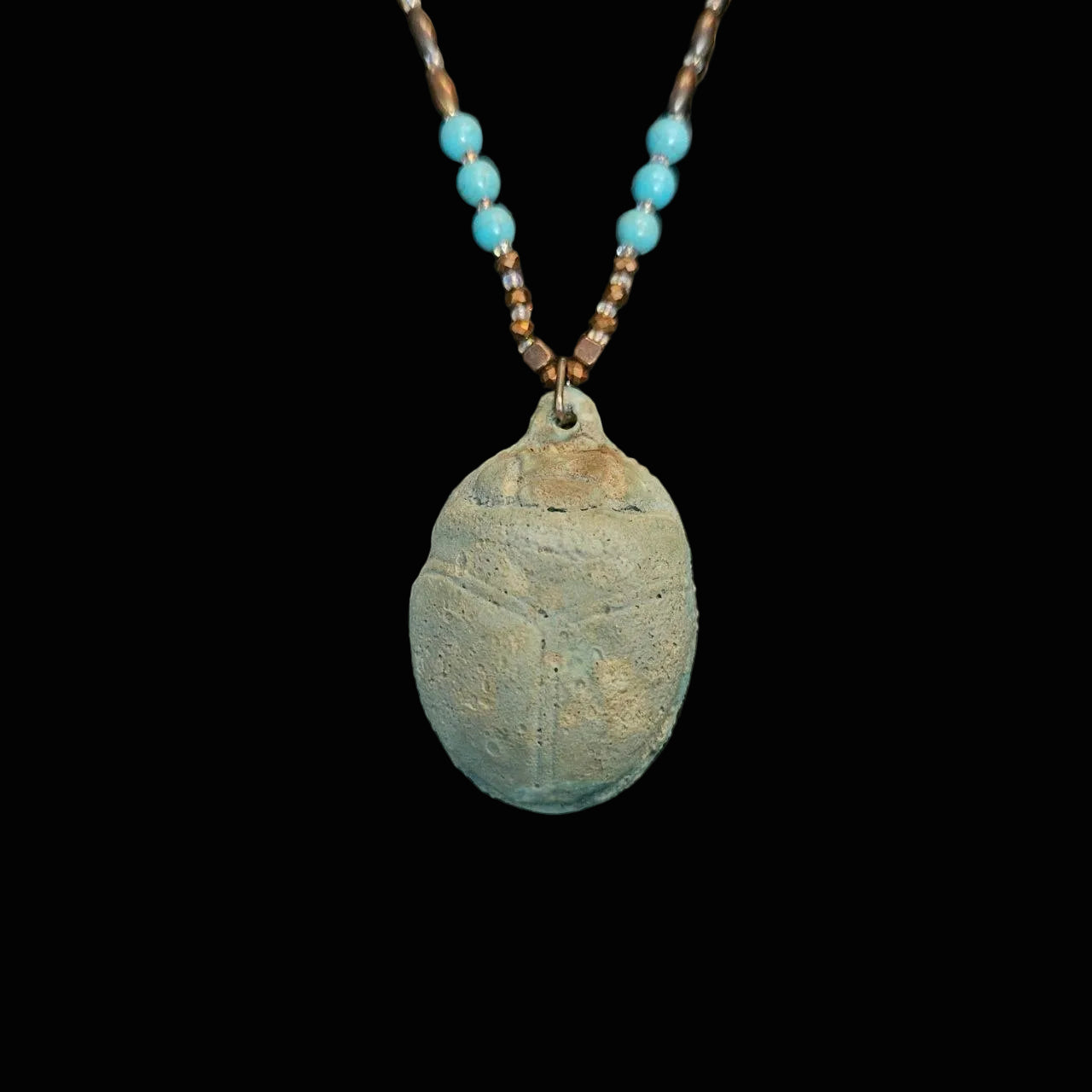 A handmade necklace featuring a textured, earthy greenish-gray beetle pendant ocarina suspended from a beaded chain with turquoise and brown beads, displayed on a dark textured background.