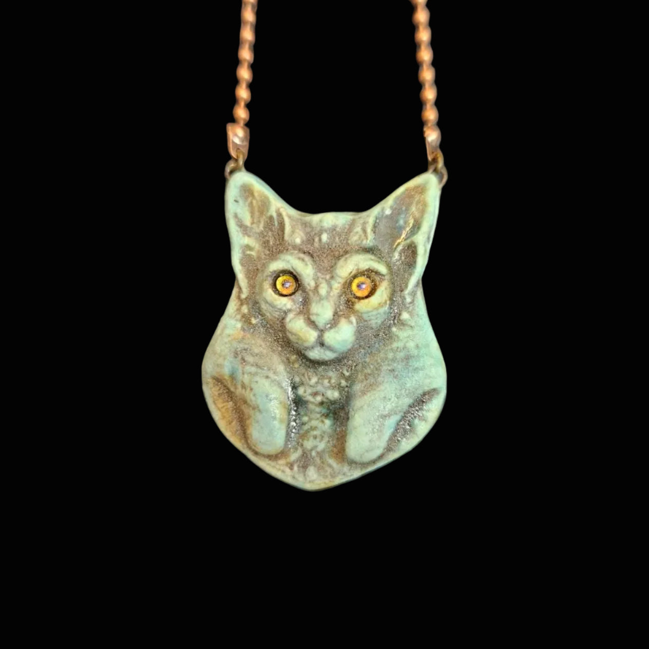 A handmade ceramic cat pendant necklace with a textured, patinated green and brown finish and orange eyes, hanging from a copper-colored beaded chain on a dark textured background.