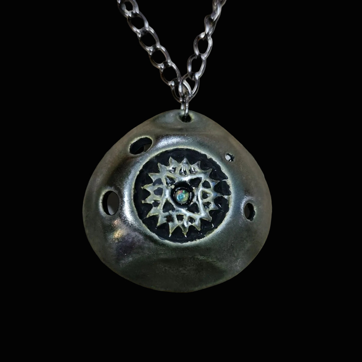 A dark metallic pendant ocarina necklace with a textured, organic shape, featuring a central starburst design and a small blue-green stone, resting on a scratched wooden surface.