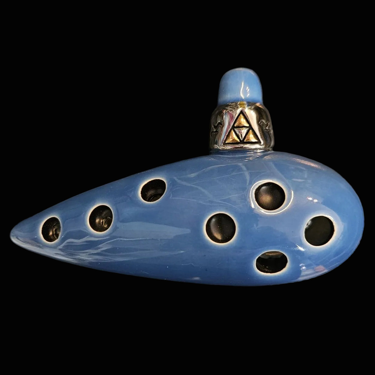 A blue ceramic ocarina with multiple finger holes and a silver metal neck featuring a gold Triforce symbol, resting on a dark textured surface.