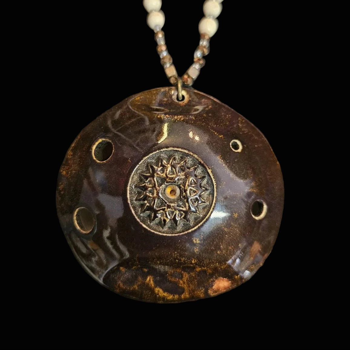 A handmade ceramic or stone pendant necklace with a dark brown marbled finish, featuring a central bronze-toned mandala medallion and a beaded chain with cream-colored beads, displayed on a dark textured background.