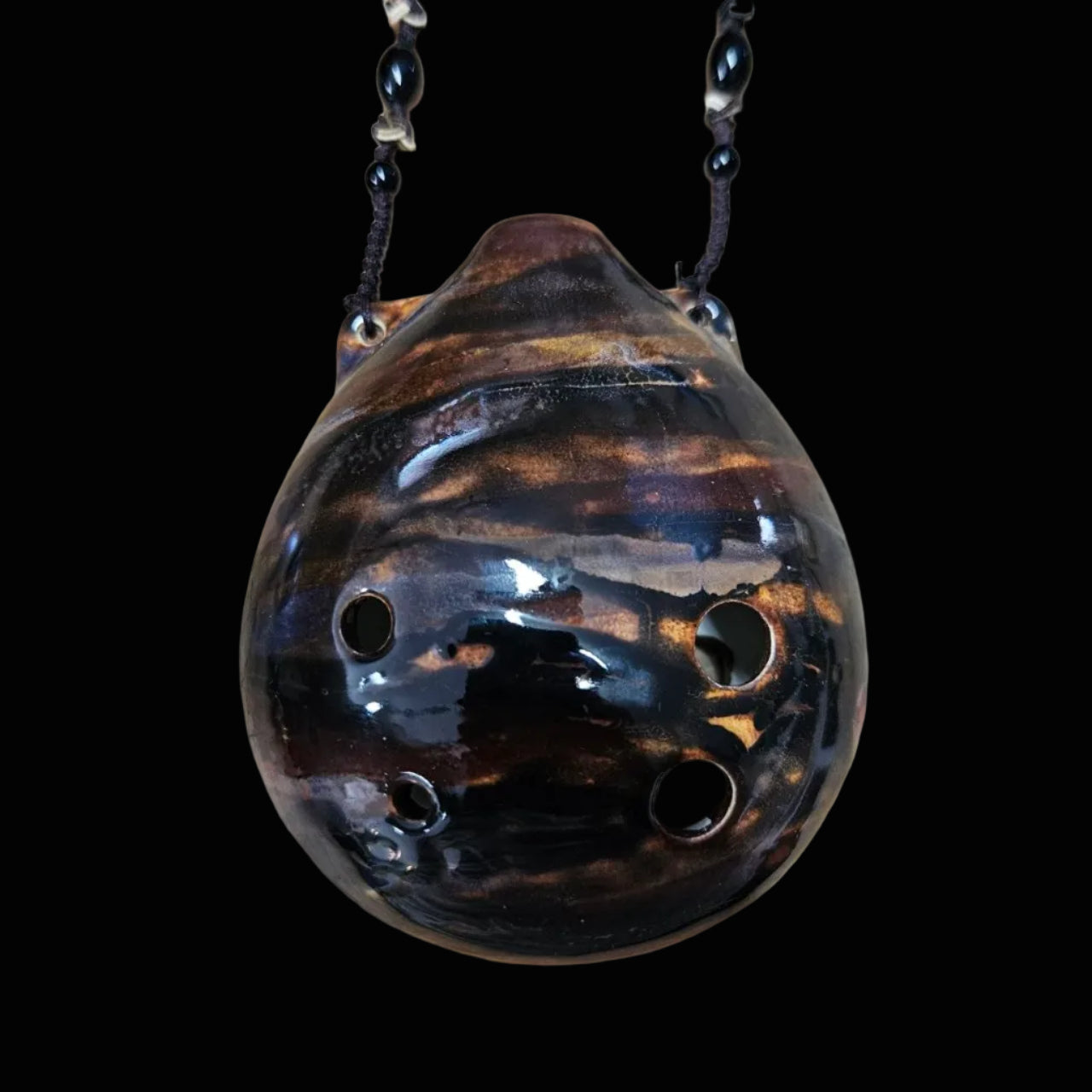 A handmade ceramic ocarina with a glossy dark brown and black marbled glaze, resting on a scratched wooden surface. Two black beaded necklaces are attached to the bottom openings.