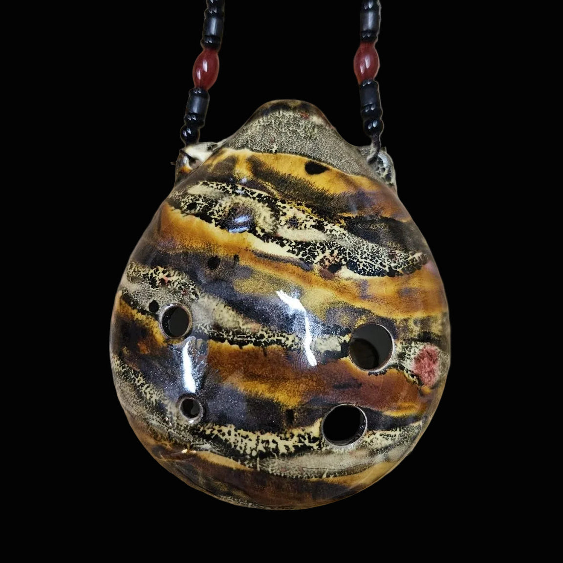 A handmade ceramic pan flute with a glossy brown, gold, and black striped glaze, resting on a scratched wooden workbench. The flute is suspended by a dark beaded necklace.
