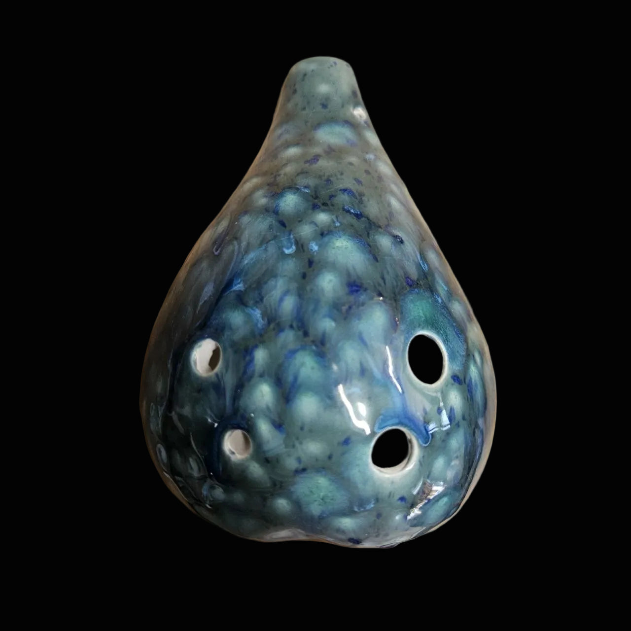 A handmade ceramic ocarina with a glossy, mottled teal and blue glaze, resting on a scratched wooden workbench.